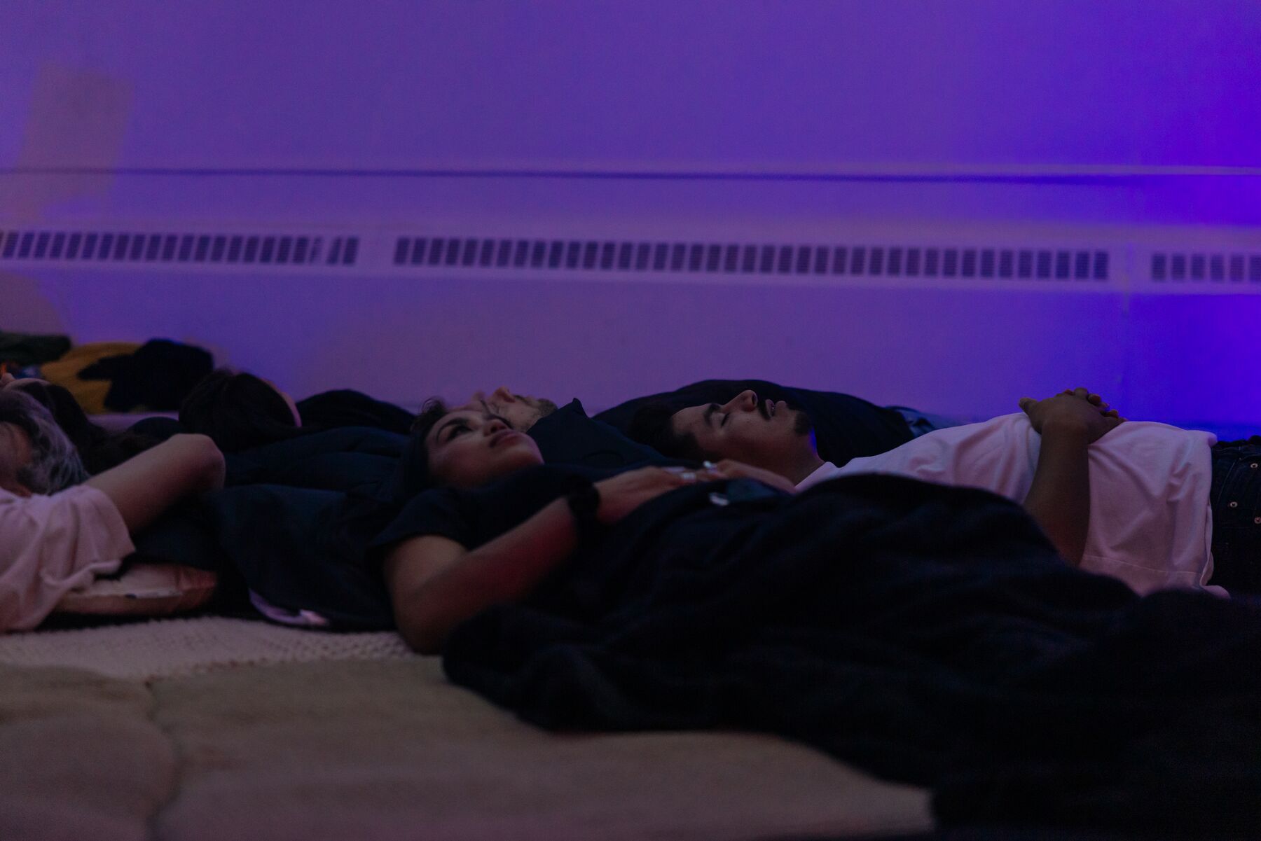 Guests lying side by side under blankets, faces up, the wall lit violet.